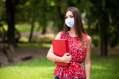 Maskeli kadın öğrenci parkta kitap tutuyor.