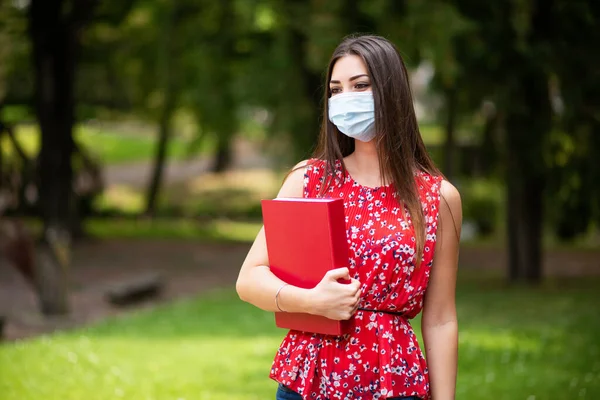 Maskeli kadın öğrenci parkta kitap tutuyor.
