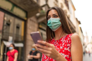 Cep telefonunu kullanarak şehirde dolaşan maskeli genç bir kadın.