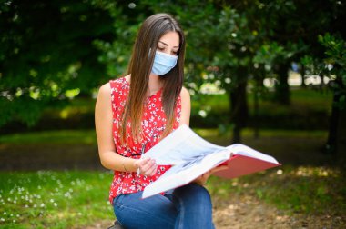 Güzel bir üniversite öğrencisi parktaki bankta kitap okuyor ve Coronavirus zamanlarında maske takıyor.