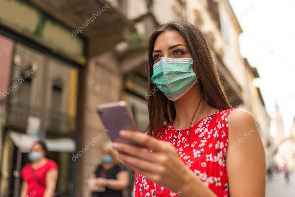 Masked young woman walking in a city while using her mobile phone