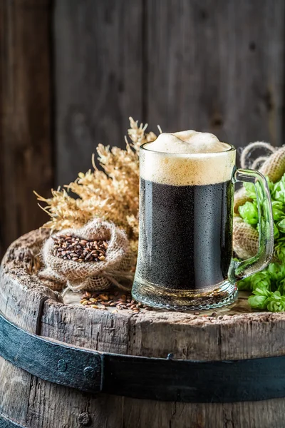Fresh pint of dark beer, hops and wheat on old barrel — Stock Photo, Image