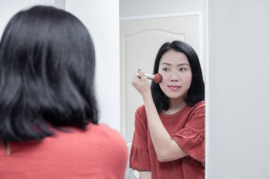 Asyalı kadın aynaya bakıyor ve allık tutarak yanak makyajı yapıyor.
