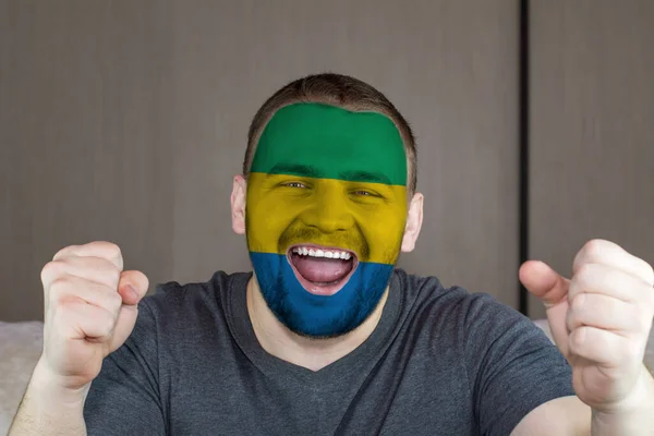 Face of young screaming man painted with flag of Gabon. Soccer team fan.