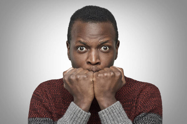 Portrait of scared young African American man dressed in sweater keeping hands in fists, holding them in front of his face, looking at camera with shocked and frightened expression. Fear concept. 