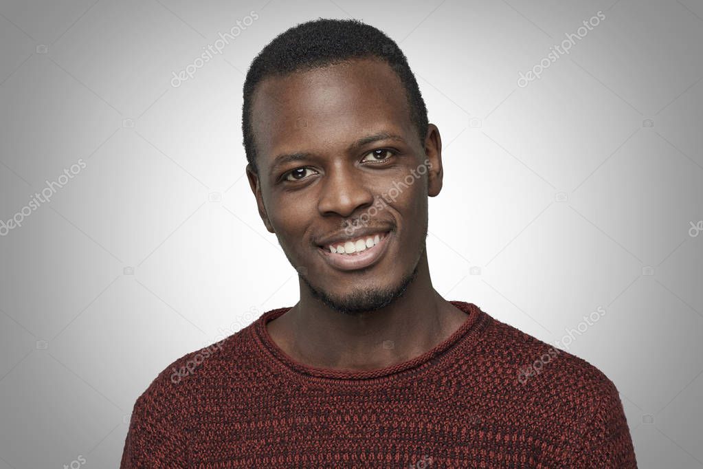 Handsome young African American male dressed in casual sweater, looking at camera with happy smile. Close up portrait of dark-skinned student smiling with his white teeth isolated on grey background