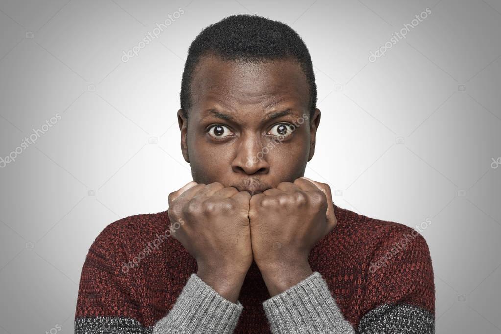 Portrait of scared young African American man dressed in sweater keeping hands in fists, holding them in front of his face, looking at camera with shocked and frightened expression. Fear concept. 