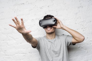 Gri t-shirt için eğlenceli, Vr Kulaklık kullanırken sanal gerçeklik yaşandığı heyecanlı yakışıklı adam beyaz tuğla duvar, dövmeli elini havada bir şey dokunmak karşı ayakta