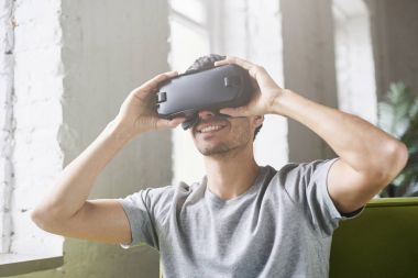 Evde yeşil kanepeye oturup video oyunu oynarken sanal gerçeklik yaşandığı oculus yarığı kulaklık, kullanan adam gülümsüyor. Avrupa erkek elele ile modern coworking Studio 3d gözlük
