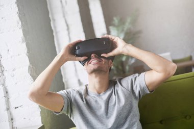 Sanal gerçeklik gözlüklü evde kanepede oturan gri t-shirt oyun giyen yakışıklı adam. Mobil oyun uygulamaları evde. Mobil apps ile yenilikçi 3d gözlük kullanma. VR teknolojisi kullanarak