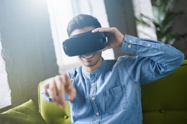 VR gözlük. Genç beyaz mavi kot gömlek erkek evde kanepede oturup video oyunu oynarken sanal gerçeklik kulaklık veya havada, parmağınızla işaret 3d gözlük gülümseyen. Lens etkisi fişekleri