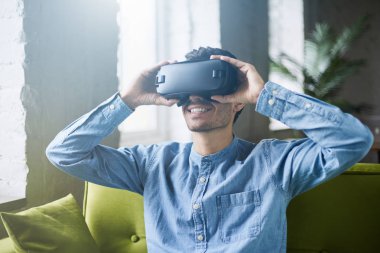 Sanal gerçeklik kulaklık ya da 3d gözlük yeşil kanepeye evde oturan mavi kot gömlek çekici genç adam. Video oyunları oynarken Vr kulaklık kullanarak yakışıklı beyaz erkek. Çok gerçekçi!
