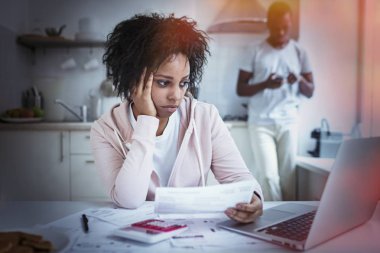 Kağıtları tutarak onun borçlarını ödemek için online banka app kullanarak onu laptop arıyorum Afrika kadın mutfak masada oturuyor. Kocası backgrond smartphone ile ayakta. Mali sorunları kavramı