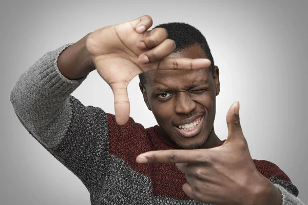 Portret van jonge African American man in warme casual trui frame met zijn handen maken, camera kijken en glimlachend, permanent tegen geïsoleerde grijze achtergrond close-up. Donkerhuidige fotograaf — Stockfoto