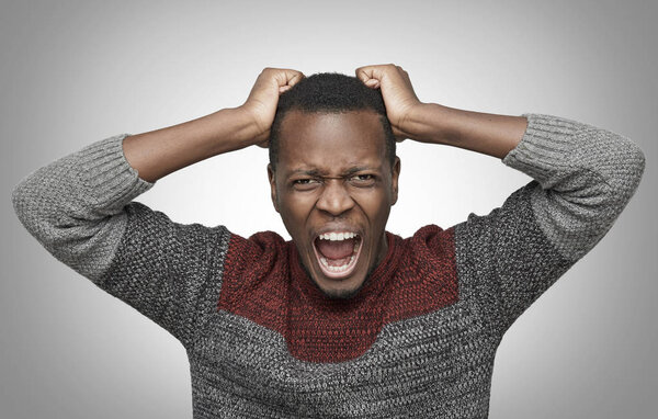 Mad African American man shouting with mouth wide open and closed eyes, holding hands on his head, trying to tear his hair out. Black male screaming with scared expression, yelling full of anger