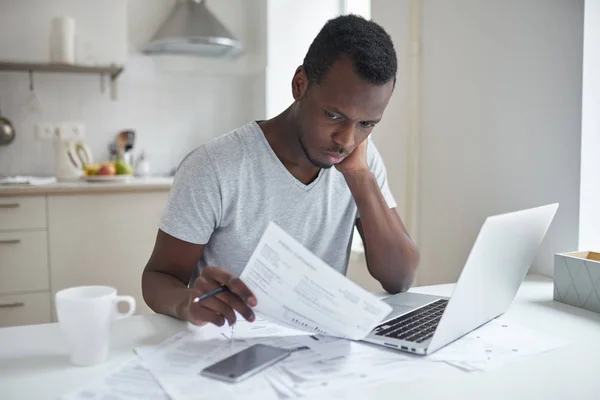 Único jovem afro-americano masculino com muitas dívidas sentindo-se estressado, calculando finanças, situando-se na mesa da cozinha com papéis, tentando fazer ambas as extremidades atender, não é capaz de pagar contas de serviços públicos — Fotografia de Stock