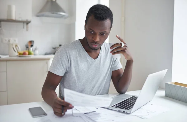 Incrível jovem afro-americano masculino, sentado à mesa da cozinha, segurando xícara de café na mão, olhando para o projeto de lei com expressão confusa concentrada, surpreso com altos impostos. Conceito de problemas financeiros — Fotografia de Stock