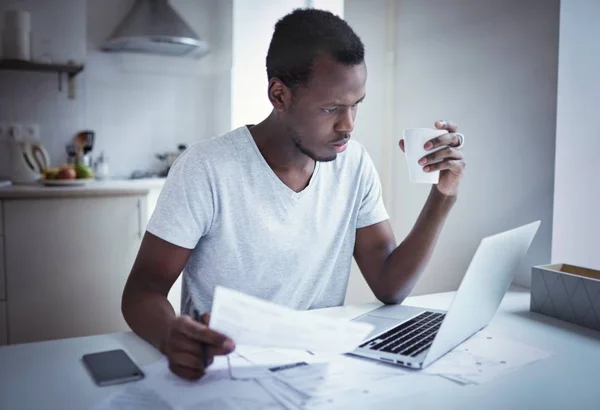 Joven hombre de piel oscura, sentado en la mesa de la cocina, sosteniendo la taza de café en la mano, mirando a la pantalla del ordenador portátil, utilizando la aplicación de banca en línea, presupuesto de planificación, cortar los gastos, tratando de ahorrar dinero —  Fotos de Stock