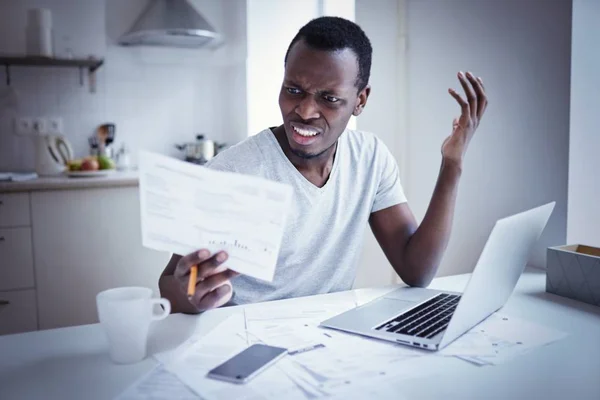 Indignant angry irritated young african american man looking at utility bill, frustrated about high taxes, worried about troubles with mortgage payment to bank, having serious problems with debt — Stock Photo, Image