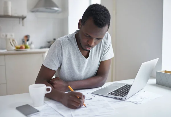 Retrato de homem de pele escura, sentado à mesa da cozinha, preenchendo formulário de inscrição, escrevendo algo em papéis, gerenciando com contas de serviços públicos, calculando despesas, tentando resolver problemas de orçamento — Fotografia de Stock