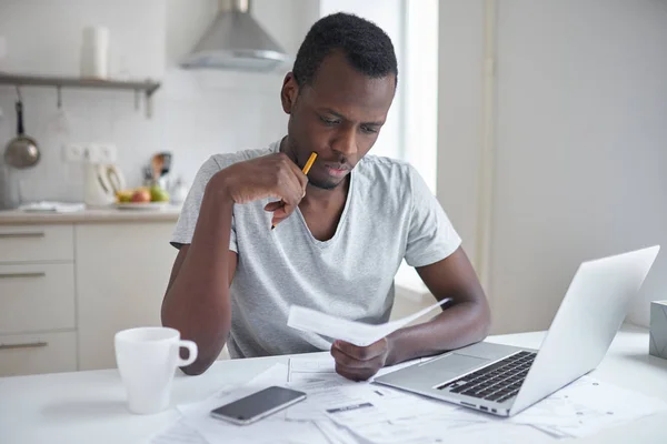 Стресс африканский американец работает через документы, расчет расходов, пытаясь сэкономить немного денег, управление финансами, сидя за кухонным столом с ноутбуком, пытаясь сделать финансовый план — стоковое фото