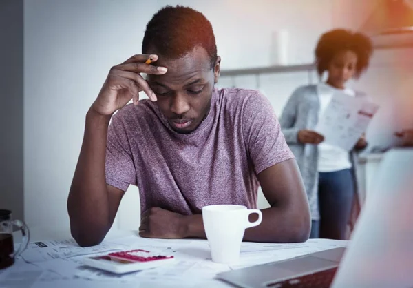 Infeliz familia afroamericana estrés financiero: hombre negro sentado en la cocina, la lectura de la notificación informando de que el banco negó a prolongar el plazo del préstamo, su esposa de pie con cuentas en segundo plano —  Fotos de Stock