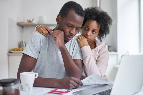 年轻的非洲裔美国人夫妇与严重的财务问题。妻子抱着失业的丈夫，而他看着试卷上表带沮丧的表情，不能还清账单 — 图库照片
