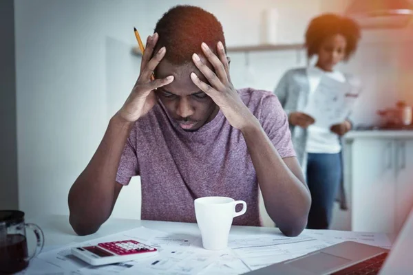 Unga afroamerikanska man sitter vid köksbordet med papper, röra huvudet med båda händerna, känsla förkrossad, inte kunna betala räkningar på grund av skulder, hans fru står i bakgrunden — Stockfoto