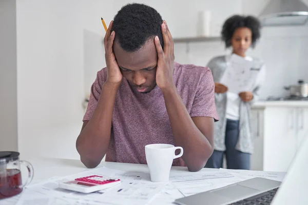 Afrikanska män sitter vid köksbordet, känner sig ledsen och deprimerad om skulder, inte kan betala sina räkningar. Hustrun står i bakgrunden med papper från banken. Människor och ekonomi koncept — Stockfoto