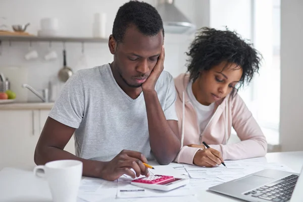 Homem africano infeliz sentado na cozinha, fazendo cálculos e sua esposa preenchendo papéis. Casal jovem ter grandes problemas ao calcular o orçamento doméstico, frustrado com a quantidade de despesas — Fotografia de Stock