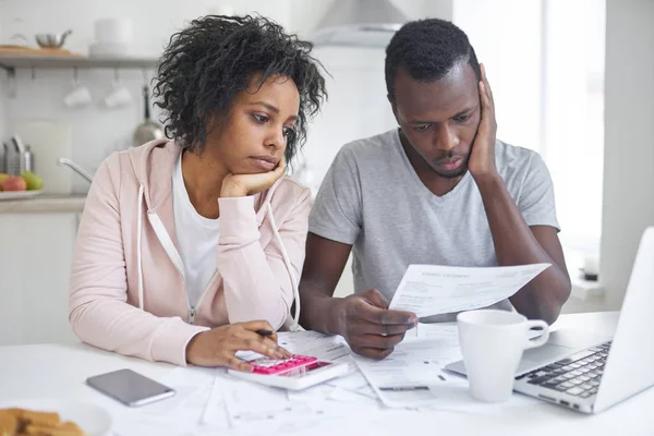 Gestresstes afrikanisch-amerikanisches Paar, das frustriert wirkt, kein Geld hat, um seine Schulden zu begleichen, das Familienbudget gemeinsam verwaltet, mit vielen Papieren, Laptop und Taschenrechner am Küchentisch sitzt — Stockfoto