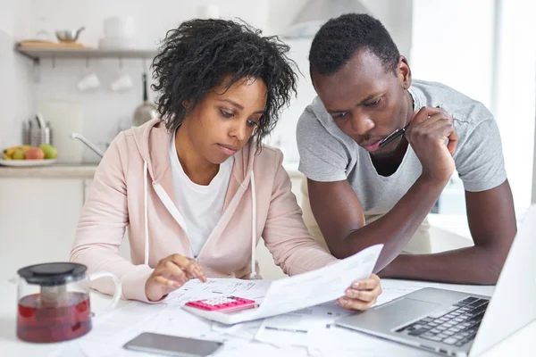 年轻的非洲裔美国夫妇在一起做文书工作，规划家庭预算，计算国内开支，与便携式计算机和计算器在家里坐在厨房的桌子。财政问题的概念 — 图库照片
