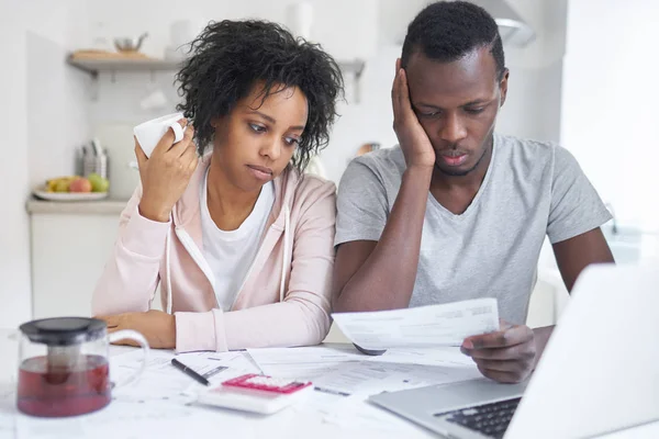 Deprimerade unga afroamerikanska paret sitter vid köksbordet med massor av papper. Familjens budget och skulder koncept. Man håller anmälan från bank, försöker beräkna inhemska kostnader — Stockfoto