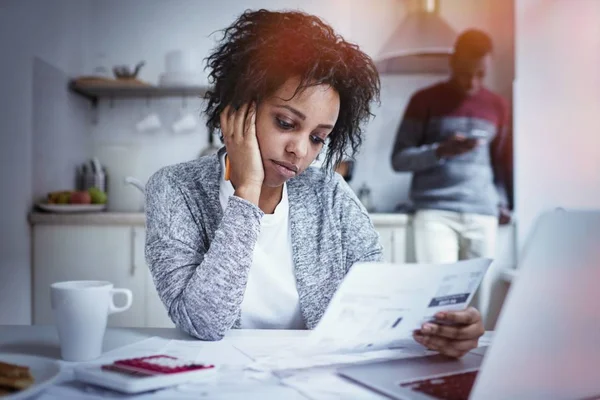 强调年轻非洲裔美国女人做文书工作在家，有沮丧的外观，同时从银行阅读通知，未能支付贷款的时间，同时在线的丈夫支票帐户余额 — 图库照片