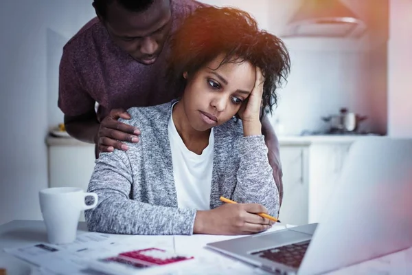 Jovem casal afro-americano com sérios problemas financeiros. Marido tentando animar a esposa triste, olhando para a tela do laptop, sentindo-se estressado ao gerenciar o orçamento da família na cozinha . — Fotografia de Stock