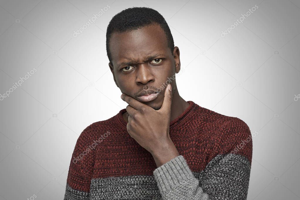 Close up portrait of serious puzzled African American male touching his chin, looking thoughtful and skeptical about something. Dark-skinned student deep in thoughts, hesitating to make decision