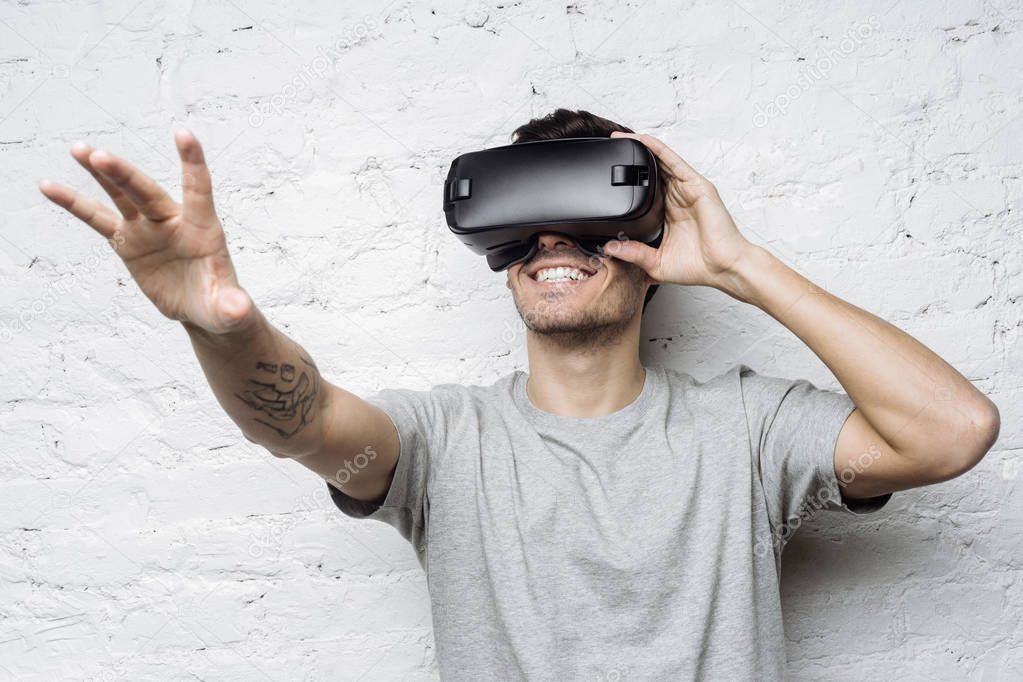Close up portrait of handsome male with tatttooed arm in grey t-shirt, experiencing virtual reality using VR headset glasses. Young man wearing goggles, playing video games against white brick wall