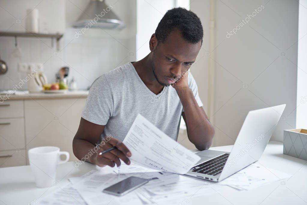 Single young african american male with many debts feeling stressed, calculating finances, siting at kitchen table with papers, trying to make both ends meet, not able to pay off utility bills