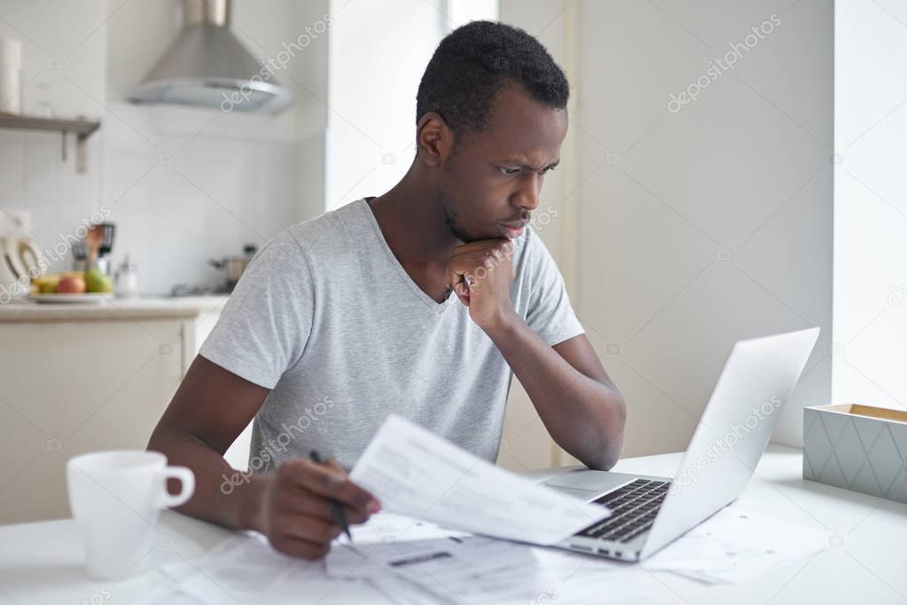 Young serious african american man sitting at kitchen table in front of open laptop, concentrated on paperwork, paying domestic bills using online banking application, planning household budget
