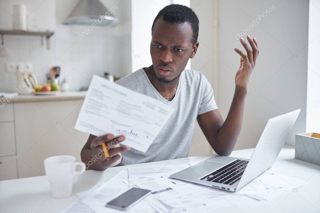 Indignant angry irritated african american male sitting at kitchen table, looking at papers in shock, astonished with amount of unpaid bills. Financial stress, feeling depressed and frustrated