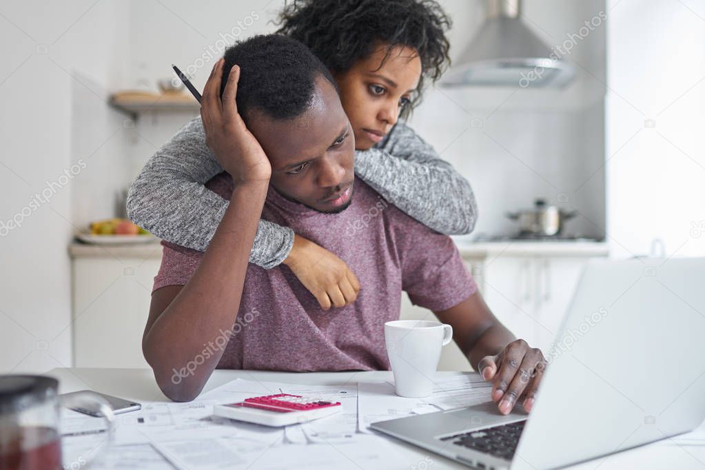 African-american female hugging her unemployed husband, while he looking at laptop, using online banking with frustrated expression, not able to pay off utility bills. Financial problems concept