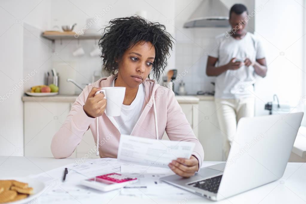 Young sad african american female drinking coffee, busy working through finances, sitting at kitchen table with open laptop, calculator and papers, her husband using mobile phone in background