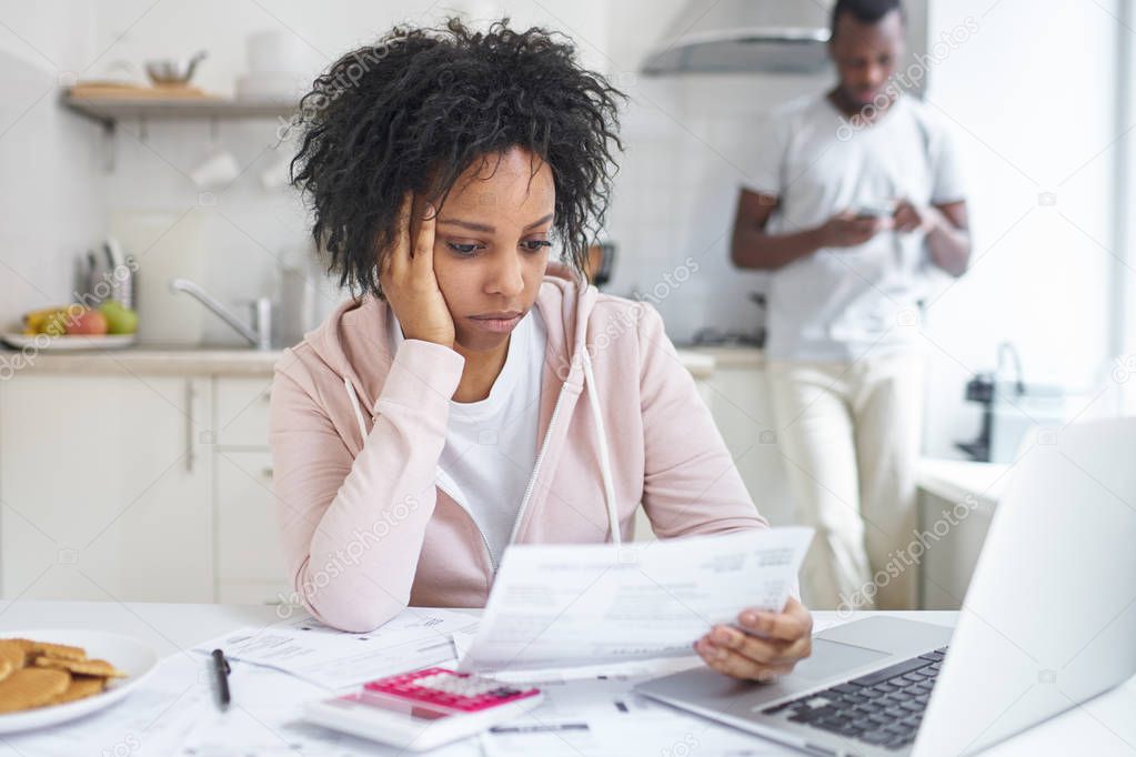 Worried african american wife sitting at kitchen table, reading notification from bank, trying to pay gas and electricity bills online, while her husband standing with smartphone on background