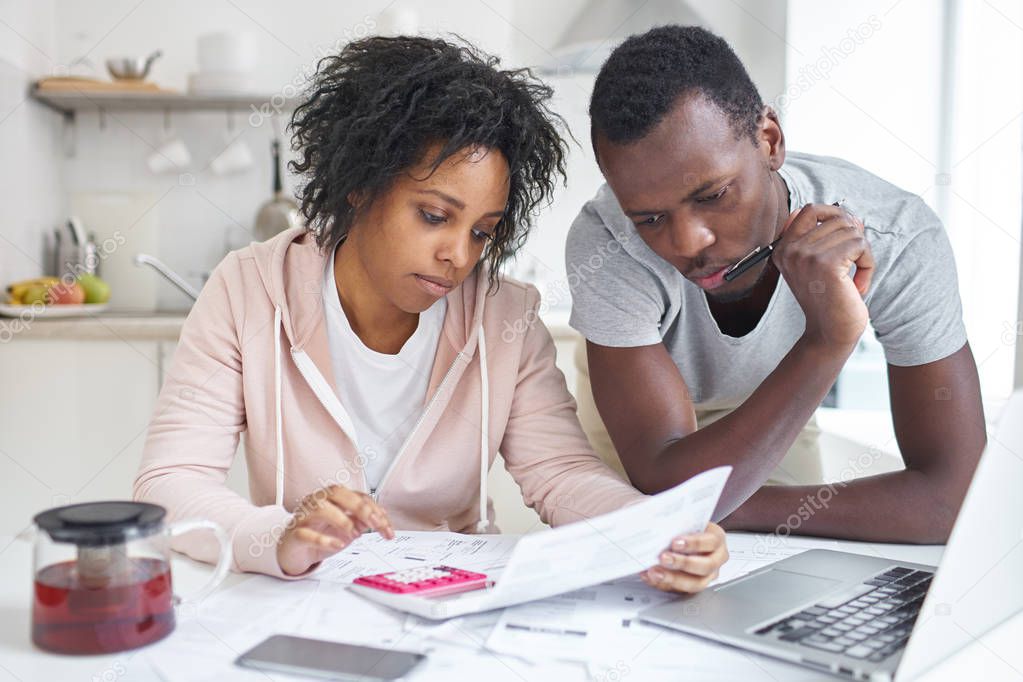 Young african american couple doing paperwork together, planning family budget, calculating domestic expenses, sitting at kitchen table with laptop and calculator at home. Financial problems concept