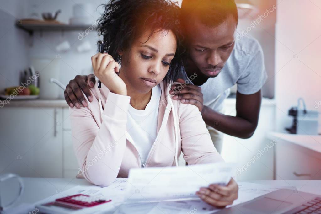 African man hugging wife, while she looking at laptop, using online banking app with, not able to pay off gas and electricity bills. Young couple sitting at kitchen table, facing financial problem 