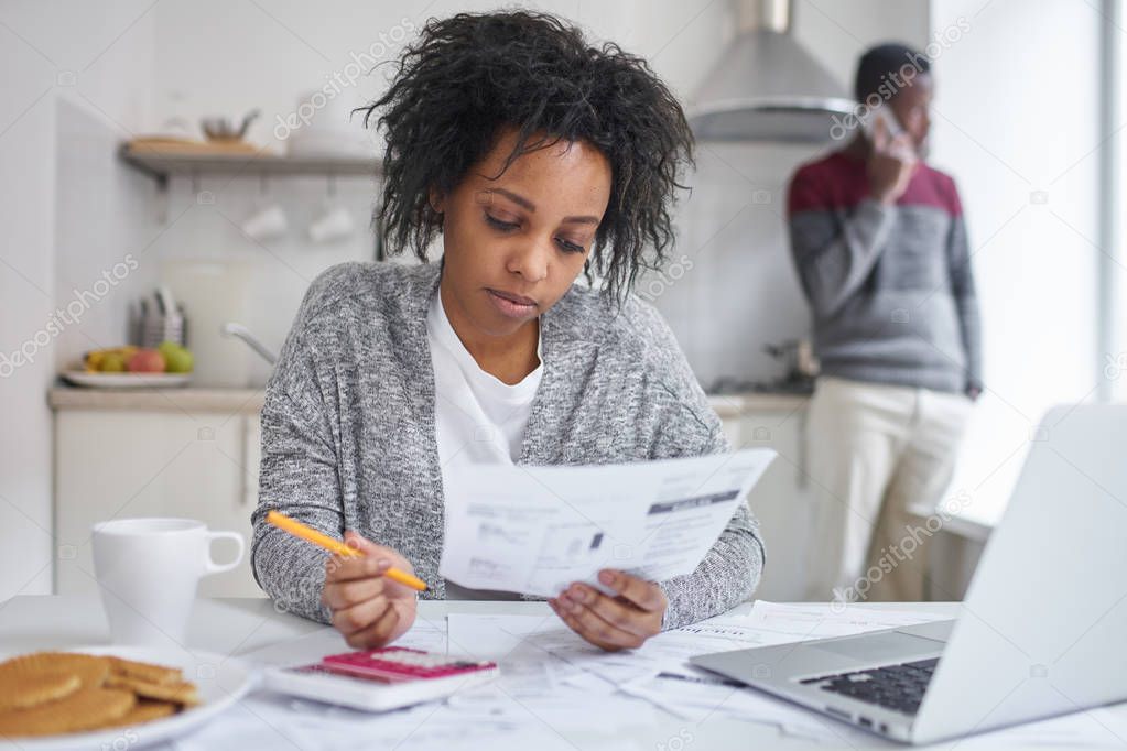 African american female reading piece of paper, calculating gas and electricity bills, sitting at kitchen table with laptop, calculator and documents, managing family finances, husband on background