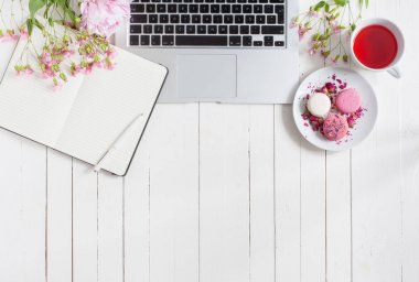 Kadınsı düz çalışma alanı laptop, defter, fincan çay, macarons ve beyaz tahta masaya çiçek ile yatıyordu. Üstten Görünüm kadar sahte.