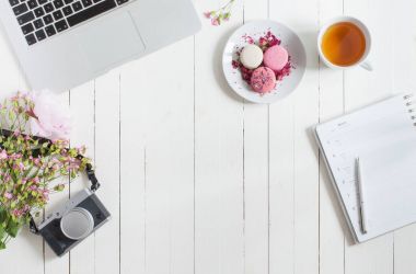 Kadınsı düz çalışma alanı laptop, fincan çay, macarons, fotoğraf makinesi ve beyaz tahta masaya çiçek ile yatıyordu. Üstten Görünüm kadar sahte.