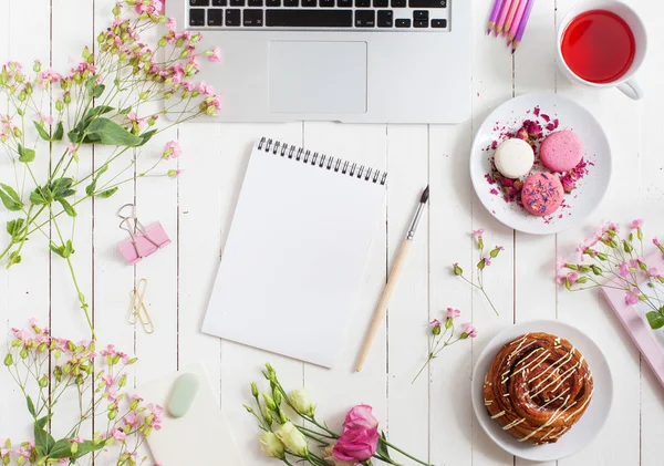 Feminina platt låg arbetsyta med pensel, anteckningsboken, laptop, kopp te, macarons och blommor på vit träbord. Ovanifrån mock upp. — Stockfoto