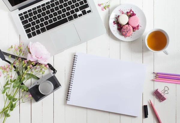 Ruang kerja rata feminin dengan laptop, secangkir teh, makaron, kamera foto dan bunga di atas meja kayu putih. tiruan tampilan atas . — Stok Foto